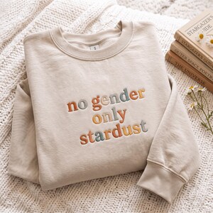 May include: A beige crewneck sweatshirt featuring the embroidered text no gender only stardust in a multi-coloured palette of muted orange, sage green, and grey. The lettering is stitched in a rounded, lowercase font across the chest area.