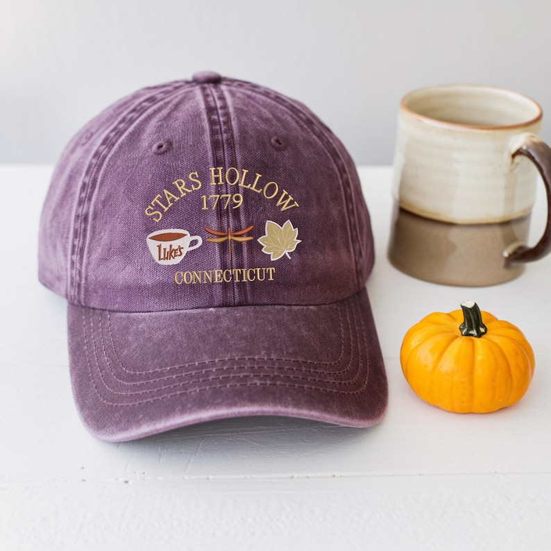 May include: A purple baseball cap with the text "Stars Hollow 1779 Connecticut" and a coffee cup, dragonfly, and leaf design. A beige and brown mug and a small orange pumpkin are also in the image.