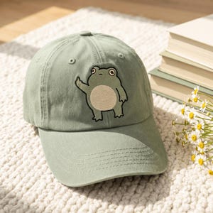 May include: A sage green baseball cap featuring an embroidered graphic of a cartoon frog with a light beige belly. The frog is depicted standing upright with one arm raised, showcasing a simple, clean design on the front panel of the hat.