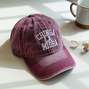 May include: A burgundy baseball cap with the text "CHINGA LA MIGRA" embroidered in white. The cap has a curved brim and a six-panel design. The hat is made of a soft, worn material.
