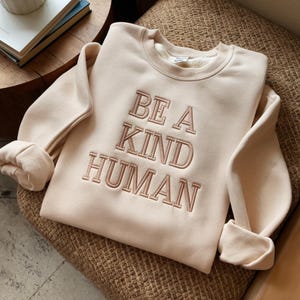 May include: A beige sweatshirt with the text "BE A KIND HUMAN" embroidered in a light brown color.
