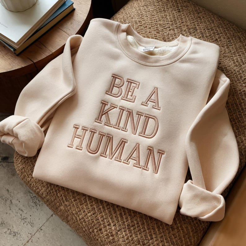May include: A beige sweatshirt with the text "BE A KIND HUMAN" embroidered in a light brown color.