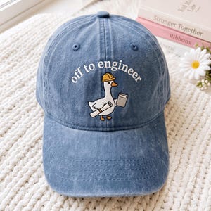 Puede incluir: Una gorra de béisbol de mezclilla azul con un bordado de un ganso blanco que lleva un casco amarillo, sostiene un plano enrollado y una tableta gris, con el texto blanco "off to engineer" arqueado sobre la ilustración del ganso.