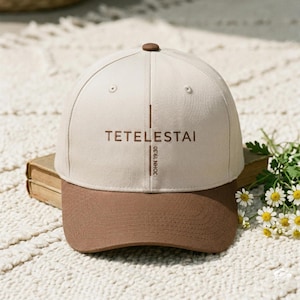 Könnte beinhalten: Beige und braune Baseballkappe mit dem Wort "TETELESTAI" auf der Vorderseite und "JOHN 19:30" vertikal auf der Seite. Die Kappe hat einen braunen Knopf oben. Ein klassisches, lässiges Accessoire.