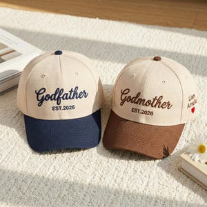 May include: Two baseball caps with the words "Godfather" and "Godmother" embroidered on the front. The caps are beige with navy blue and brown brims, respectively. The text "EST.2026" is also embroidered on the front.