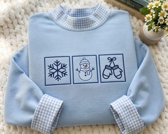 May include: A light blue long-sleeve sweatshirt featuring a mock neck and cuffs with a blue and white gingham pattern. The front displays three embroidered square panels containing a snowflake, a snowman, and a pair of mittens.
