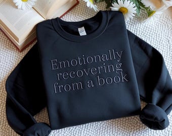 Puede incluir: Sudadera negra con el texto bordado "Emotionally recovering from a book". La sudadera de cuello redondo está doblada sobre una superficie blanca, con un libro y margaritas en el fondo.