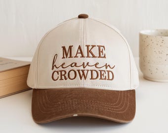 May include: A cream-coloured baseball cap with a dark brown brim and a dark brown button on top. The front of the cap features the embroidered text "MAKE heaven CROWDED" in a dark brown font, combining block letters and cursive script.