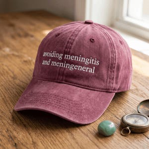 May include: A maroon baseball cap with a six-panel construction and a curved brim. The front of the cap features white embroidered text that reads: avoiding meningitis and meningeneral. The fabric has a slightly faded, washed-out appearance.
