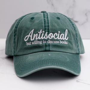 May include: A green baseball cap with the text "Antisocial but willing to discuss books" embroidered in white.