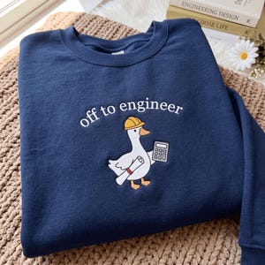Könnte beinhalten: Ein marineblaues Sweatshirt mit Rundhalsausschnitt und einer Stickerei, die eine weiße Gans mit gelbem Schutzhelm zeigt, welche einen Taschenrechner und eine zusammengerollte Blaupause hält, darüber der weiße Schriftzug "off to engineer".