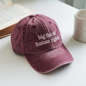 May include: A maroon baseball cap with the text "big fan of human rights" in white. The cap is made of a soft, worn material and has a curved brim. The hat is sitting on a white surface.
