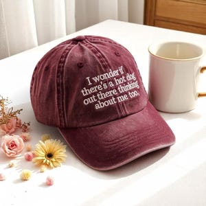 May include: A maroon baseball cap with the embroidered text "I wonder if there's a hot dog out there thinking about me too." A white mug with a gold rim is next to the cap. The cap is on a white surface.