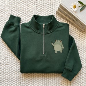 May include: A dark green quarter-zip sweatshirt featuring a small graphic of a light green frog on the left chest. The frog is depicted in a standing position with one arm raised. The sweatshirt has a high collar with a silver metal zipper.