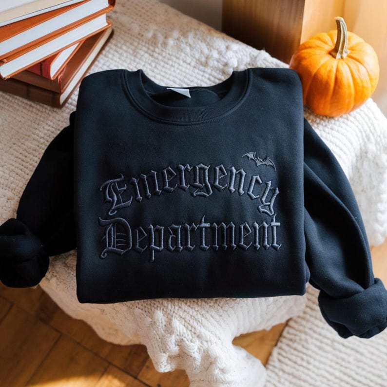 May include: A black sweatshirt with the words "Emergency Department" embroidered in a gothic font, with a small bat above the text. The sweatshirt is folded on a white, textured surface, with a small pumpkin and a stack of books in the background.