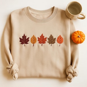 May include: A beige sweatshirt featuring embroidered autumn leaves in shades of red, orange, and brown. A small orange pumpkin and a beige mug are placed on the right side of the sweatshirt.