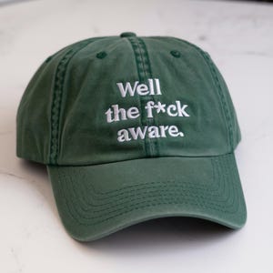 May include: A green baseball cap with white embroidered text that reads "Well the f*ck aware."