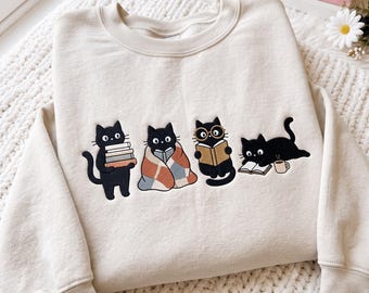 May include: A cream-colored crewneck sweatshirt featuring an embroidered graphic of four black cats. One cat holds a stack of books, one is wrapped in a plaid blanket reading, one wears round glasses reading, and one lies down with a book and mug.