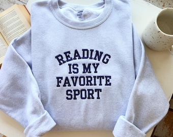 May include: A light gray sweatshirt with the text "READING IS MY FAVORITE SPORT" in navy blue. The sweatshirt has a crew neck and long sleeves, with the sleeves cuffed at the wrists. A book and a mug are in the background.