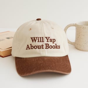 May include: A beige baseball cap with a brown brim and the embroidered text "Will Yap About Books" in brown. The cap has a curved brim and a button on top. A white mug and a book are in the background.