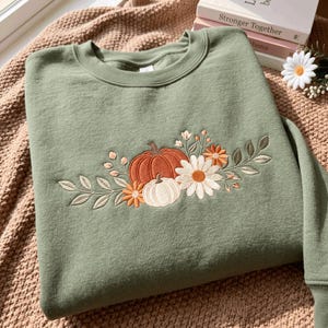 May include: A sage green crewneck sweatshirt featuring an embroidered design on the chest. The embroidery depicts a large orange pumpkin, a smaller white pumpkin, white daisies, orange flowers, and green leafy branches extending to the sides.
