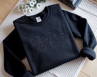 May include: A black sweatshirt with the words "DENTAL HYGIENIST" embroidered across the front. The sweatshirt is displayed on a cream-colored, textured surface, with a sprig of white daisies and a stack of books in the background.