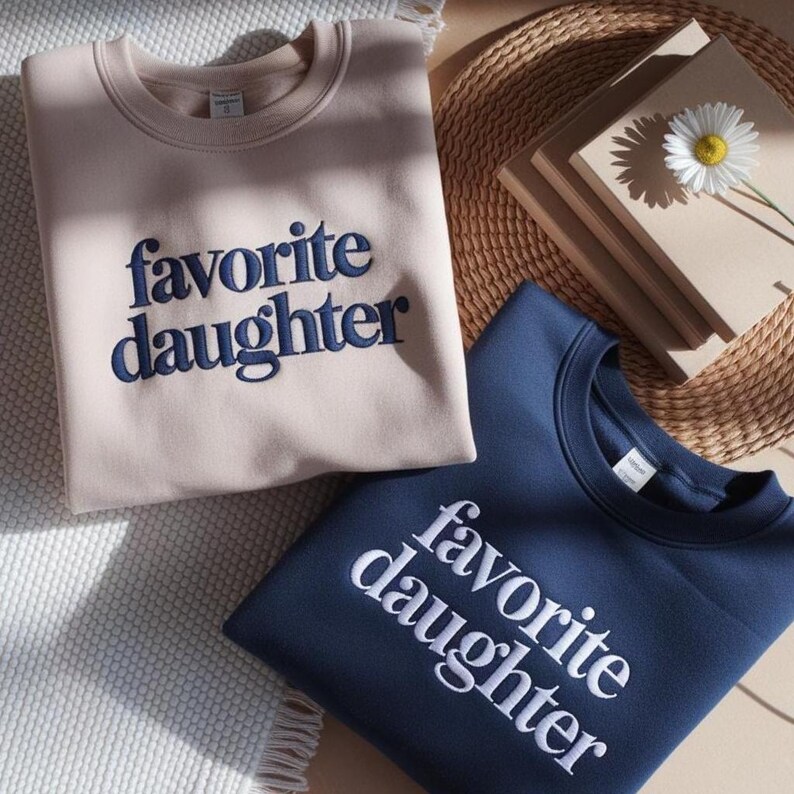 May include: Two crewneck sweatshirts, one tan and one navy blue, embroidered with the words "favorite daughter." The tan sweatshirt has navy blue embroidery, while the navy blue sweatshirt has white embroidery. The sweatshirts are folded and placed on a textured surface.