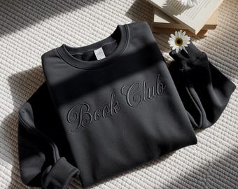 Puede incluir: Una sudadera negra de cuello redondo con el texto bordado "Book Club" en cursiva. La sudadera está sobre una superficie texturizada blanquecina, con sombras de sol. Libros y margaritas en el fondo.