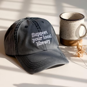 May include: A gray baseball cap with the embroidered text "Support your local library" in white. The cap is next to a speckled ceramic mug with a cork base. A dried flower is also present.