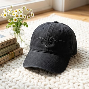 May include: A black baseball cap with a distressed finish featuring the embroidered text "auntie" in a cursive script, followed by "EST. 2026" and a small heart icon centered on the front panel of the hat.