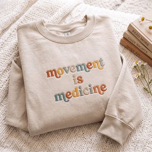May include: A beige crewneck sweatshirt featuring the embroidered text "movement is medicine" in a multi-colored font with shades of orange, teal, and yellow. The garment has long sleeves, a ribbed collar, and ribbed cuffs, showcasing a clean design.
