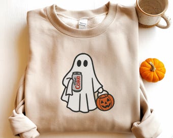 Op de afbeelding: Beige sweatshirt met een witte spookafbeelding met een Diet Coke-blikje en een pompoen-lantaarn. De geest heeft zwarte ogen en een zwarte omtrek. Een kleine oranje pompoen en een keramische mok zijn ook zichtbaar.