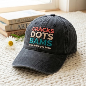 May include: A dark gray baseball cap with the words "CRACKS DOTS BAMS" in red, cream, and teal embroidery. The text "If you know, you know" is embroidered below. The cap is sitting on a white, textured surface.