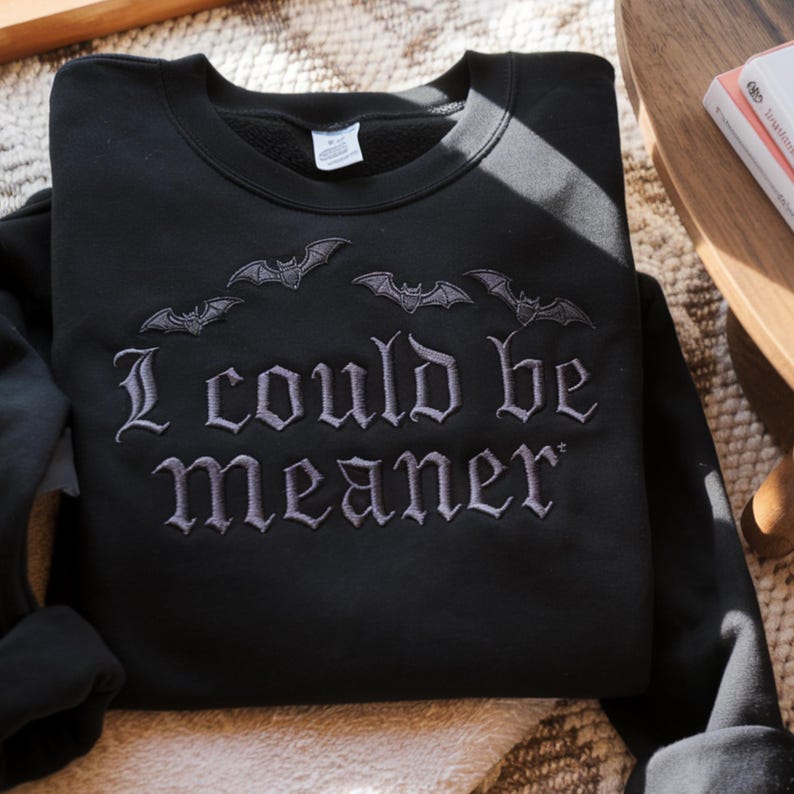 Puede incluir: Sudadera negra con murci&eacute;lagos bordados y la frase "I COULD BE MEANER" en letras g&oacute;ticas grises. Una prenda c&oacute;moda con tem&aacute;tica oscura.