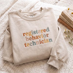 May include: A beige crewneck sweatshirt featuring the embroidered text "registered behavior technician" in a multi-colored palette of muted orange, sage green, and grey. The lettering is arranged in three lines across the center of the chest.