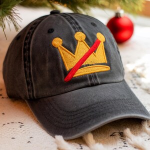 May include: A charcoal gray baseball cap with a gold crown design crossed out by a red line. The crown is embroidered on the front of the cap. The cap has a curved brim and a fabric strap closure.