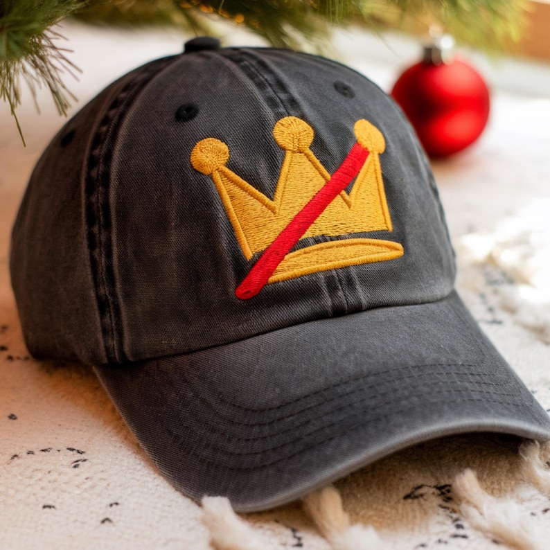 May include: A charcoal gray baseball cap with a gold crown design crossed out by a red line. The crown is embroidered on the front of the cap. The cap has a curved brim and a fabric strap closure.