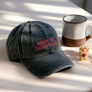 May include: A dark gray baseball cap with the embroidered text "MADE BY IMMIGRANTS" in red. The cap is slightly distressed and has a curved brim. A ceramic mug and dried flowers are in the background.