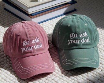Könnte beinhalten: Zwei Baseballkappen, eine in Staubrosa und eine in Salbeigrün, mit dem weißen Text "go ask your dad" bestickt. Sie liegen auf einer strukturierten weißen Oberfläche, mit Büchern und einer Gänseblümchen im Hintergrund.