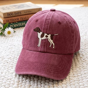 Può includere: Un cappellino da baseball color marrone scuro con un ricamo raffigurante un cane pointer bianco e marrone di profilo. Il cappello presenta una corona strutturata, una visiera curva e occhielli cuciti sui pannelli superiori.