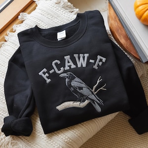 May include: Black sweatshirt with the embroidered text "F-CAW-F" above a detailed illustration of a raven perched on a branch. The raven is in shades of gray, and the branch is a light brown color.
