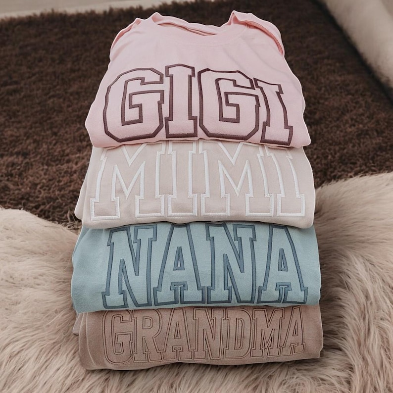 May include: Stack of four pink, beige, blue, and brown shirts with the words "Gigi", "Mimi", "Nana", and "Grandma" embroidered on them in a varsity style font.