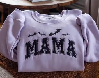 May include: A lilac sweatshirt featuring the word "MAMA" in black, block lettering, with black bat silhouettes above. The sweatshirt is folded on a brown surface.