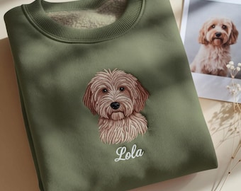 May include: Olive green sweatshirt with a detailed embroidered image of a dog's face. The dog's name, Lola, is embroidered below the image in white script. A photograph of the dog is in the background.