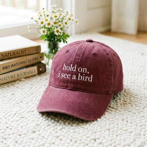 May include: A maroon baseball cap with the text "hold on, i see a bird" in white. The cap is made of a soft material and has a curved brim. The background includes books and a vase of flowers.