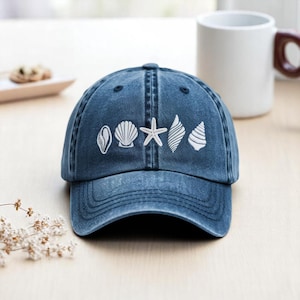 May include: A blue denim baseball cap with white embroidered seashell designs. The cap features a curved brim and an adjustable strap. The design includes a scallop shell, starfish, and other sea-themed elements.