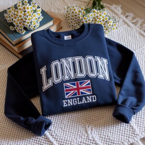 May include: Navy blue sweatshirt with the word "LONDON" in white block letters above a Union Jack flag and the word "ENGLAND" below. The sweatshirt is folded on a white textured surface with books and flowers in the background.