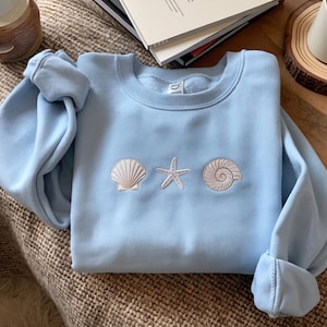 May include: A light blue crewneck sweatshirt featuring three embroidered white designs centred on the chest: a fan-shaped scallop shell, a five-pointed starfish, and a spiral seashell. The garment has long sleeves and a ribbed neckline.