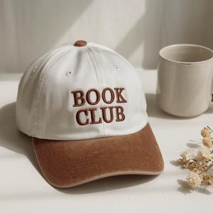 Pu&ograve; includere: Un cappellino da baseball bianco con visiera marrone e la scritta ricamata "BOOK CLUB" in marrone. Il cappellino ha un bottone marrone sulla parte superiore. Una tazza beige &egrave; sullo sfondo.