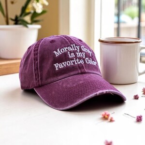 May include: A distressed purple baseball cap with the embroidered text "Morally Grey is my Favorite Color" in white. The cap is sitting on a white surface next to a white mug with a brown rim.